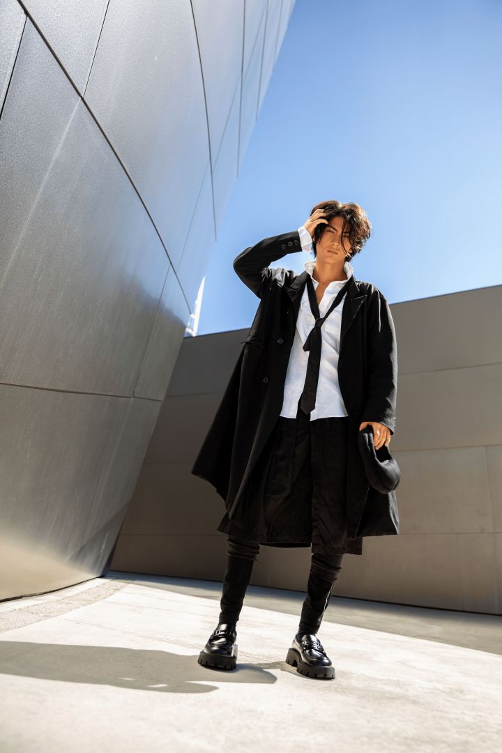 male model standing with silver shiny symmtrical walls surrounded, clothed in a black long jacket, white top with black tie. He held black, circular thicked clutch, wearing a black kilt with high platform boots.