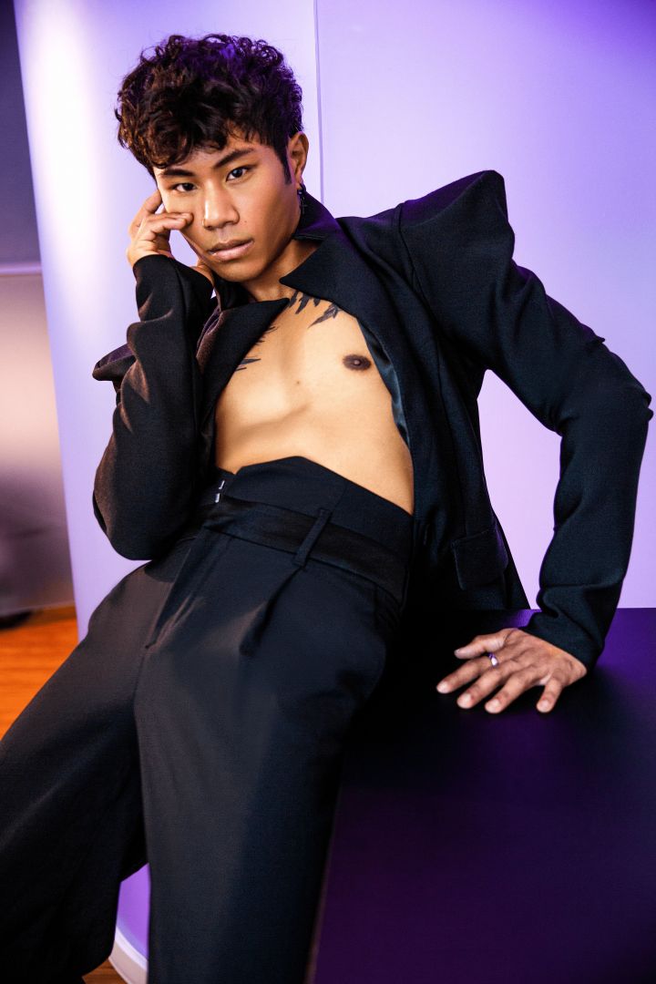 Asian male model leaning against a table, framed by a futuristic purple neon-lit backdrop. He sensually touches his face, dressed in a pointed shoulder jacket revealing his chest, paired with high-waisted pants.