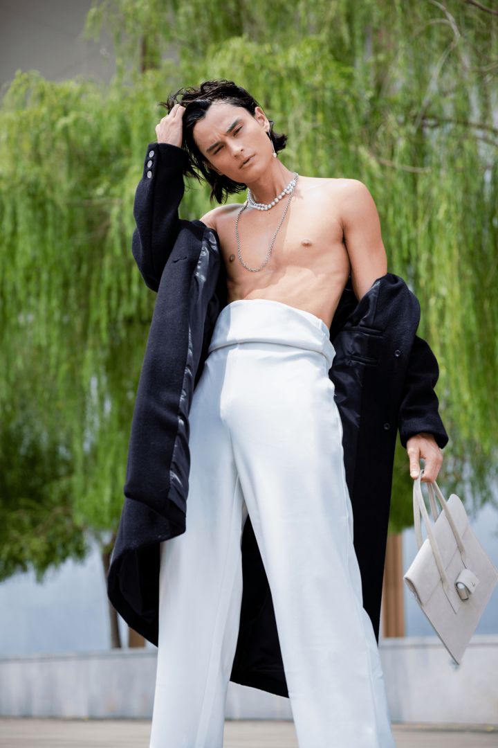 Asian male model standing in front of a willow tree and cement walls, one hand running through his hair, leaning back with jacket slipping off the shoulder, wearing high-waisted white pants and holding a bag.