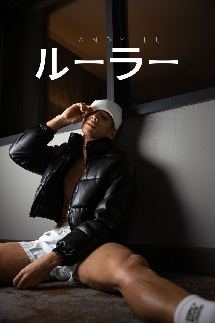 Asian male model reclining against a wall near a window, adjusting white cap over his eyes. He is dressed in a black fluffy jacket, white boxers, and socks, with one arm resting lazily on the floor between his legs.